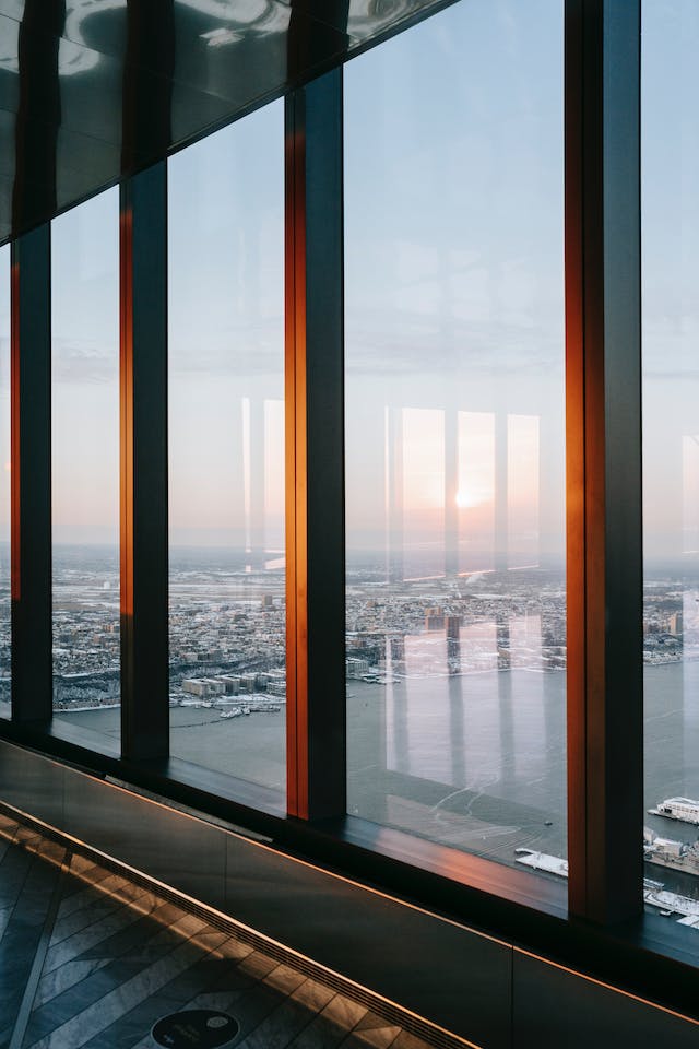 View of a coastal city at sunset through tall vertical bay windows, with soft light casting warm hues on the sleek interior floor.